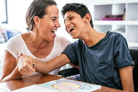 A smiling caregiver and a laughing teenage boy sitting together at a table