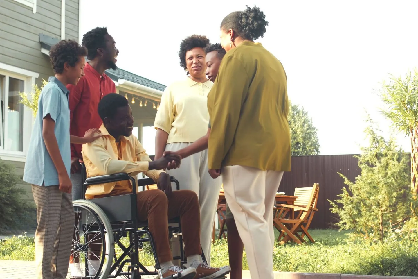 A family gathered outside a house, greeting a relative who uses a wheelchair